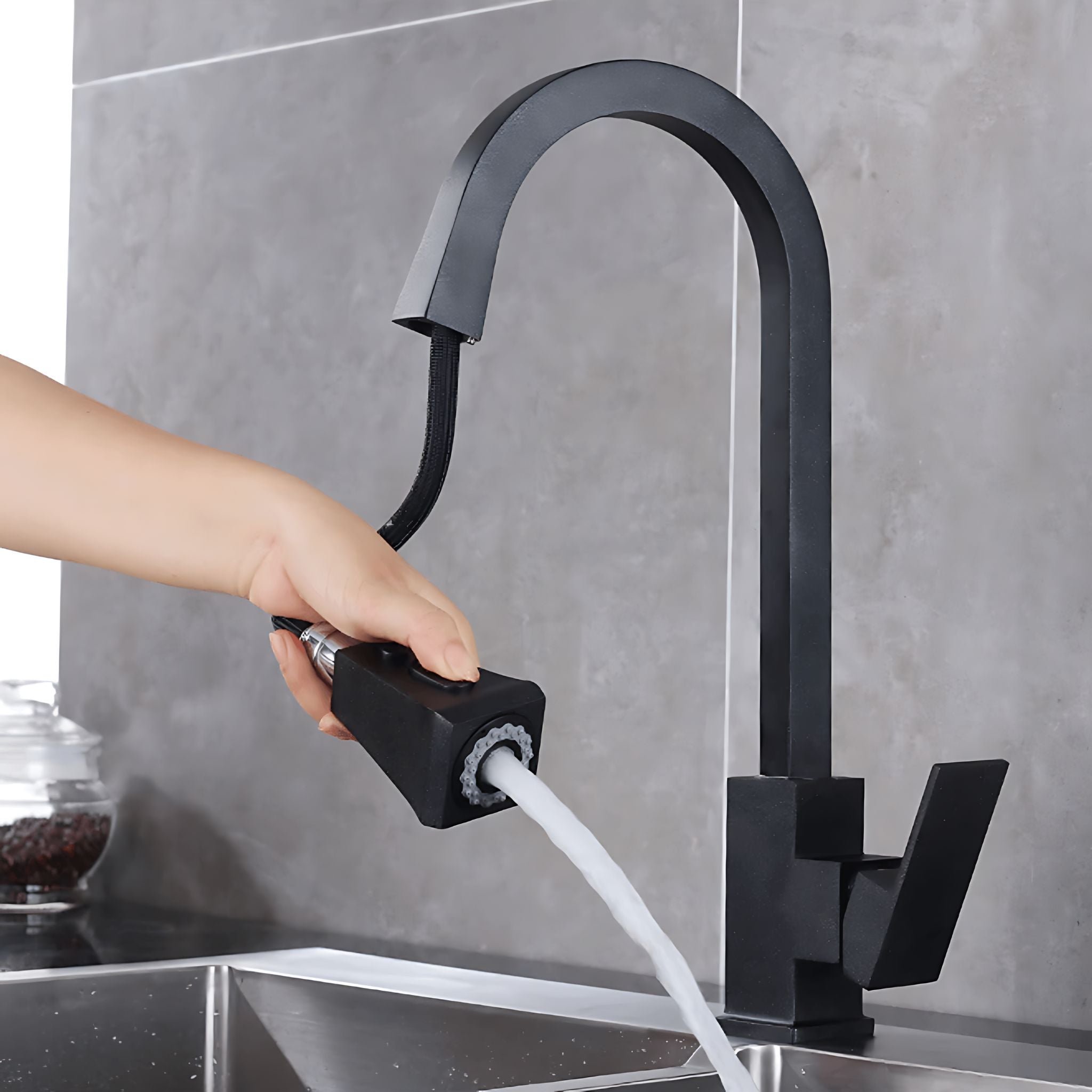 A matte black gooseneck faucet with a hand holding a matching black spray attachment under running water, set against a gray tiled kitchen backsplash. #color_matte black