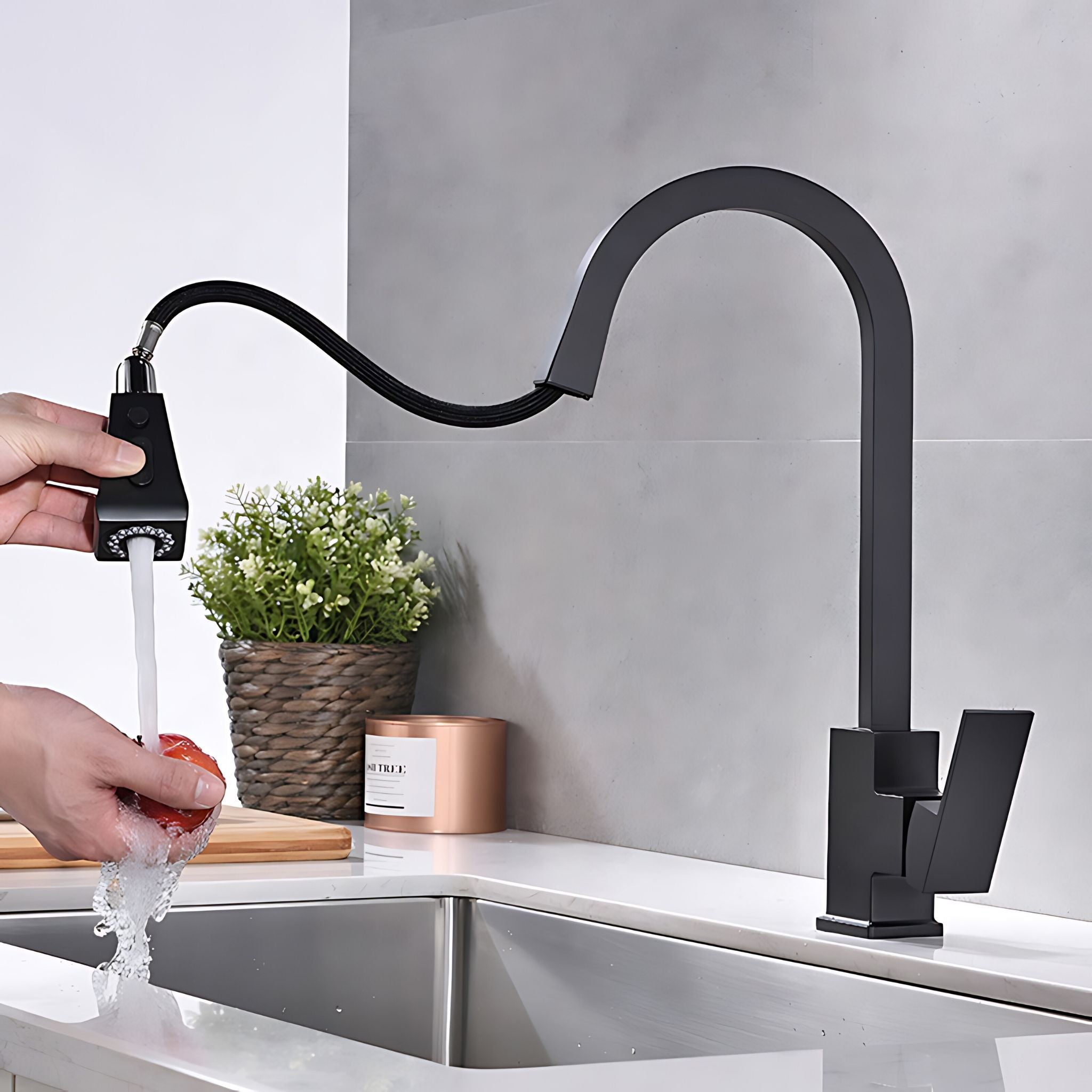 A matte black gooseneck faucet with a pull-out sprayer is shown in a kitchen setting, with a hand turning on the water while washing a red tomato. The faucet is mounted on a white countertop next to a stainless steel sink, with a gray tiled backsplash and a small potted plant in the background. #color_matte black
