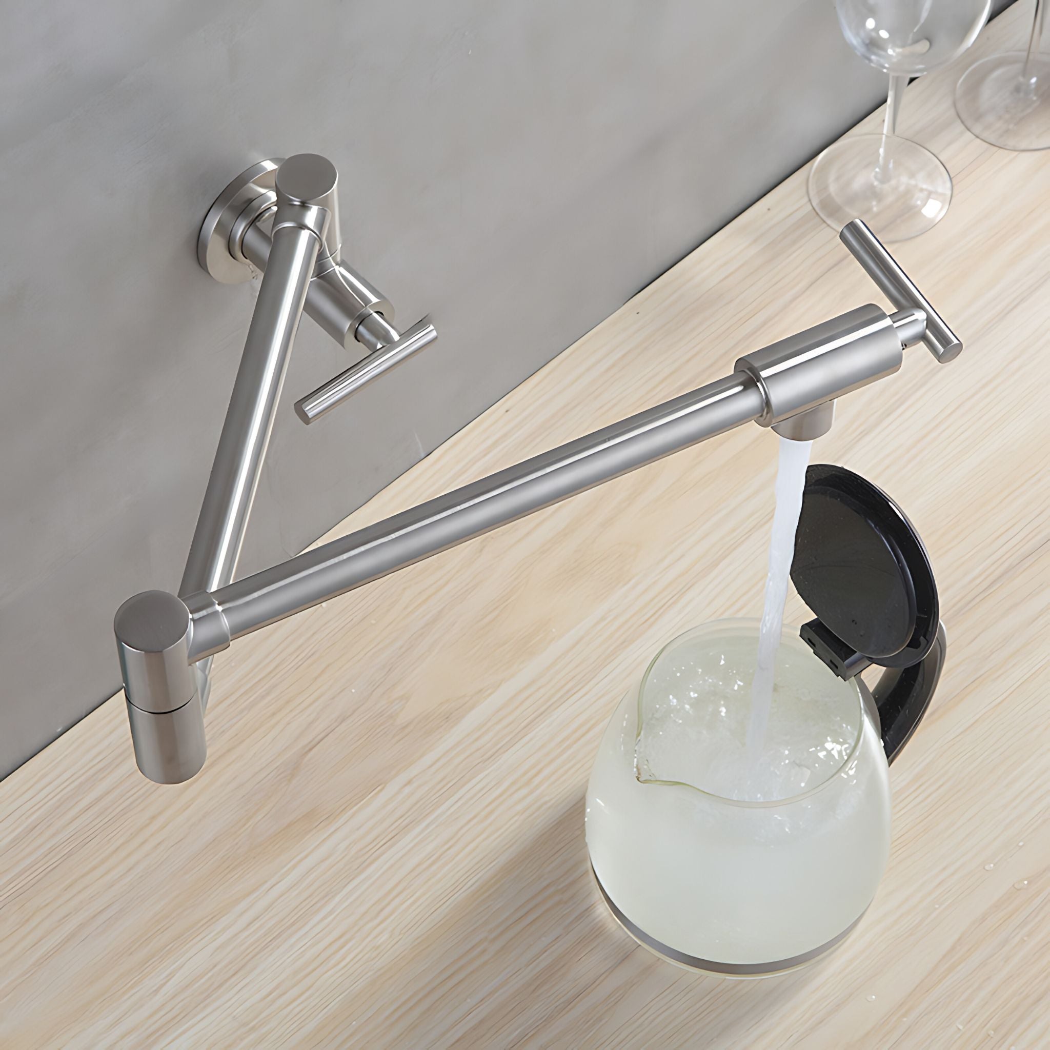 A modern wall-mounted stainless steel kitchen faucet with an adjustable arm and T-shaped handle, shown dispensing water into a glass carafe on a light wood countertop. #color_brushed nickel