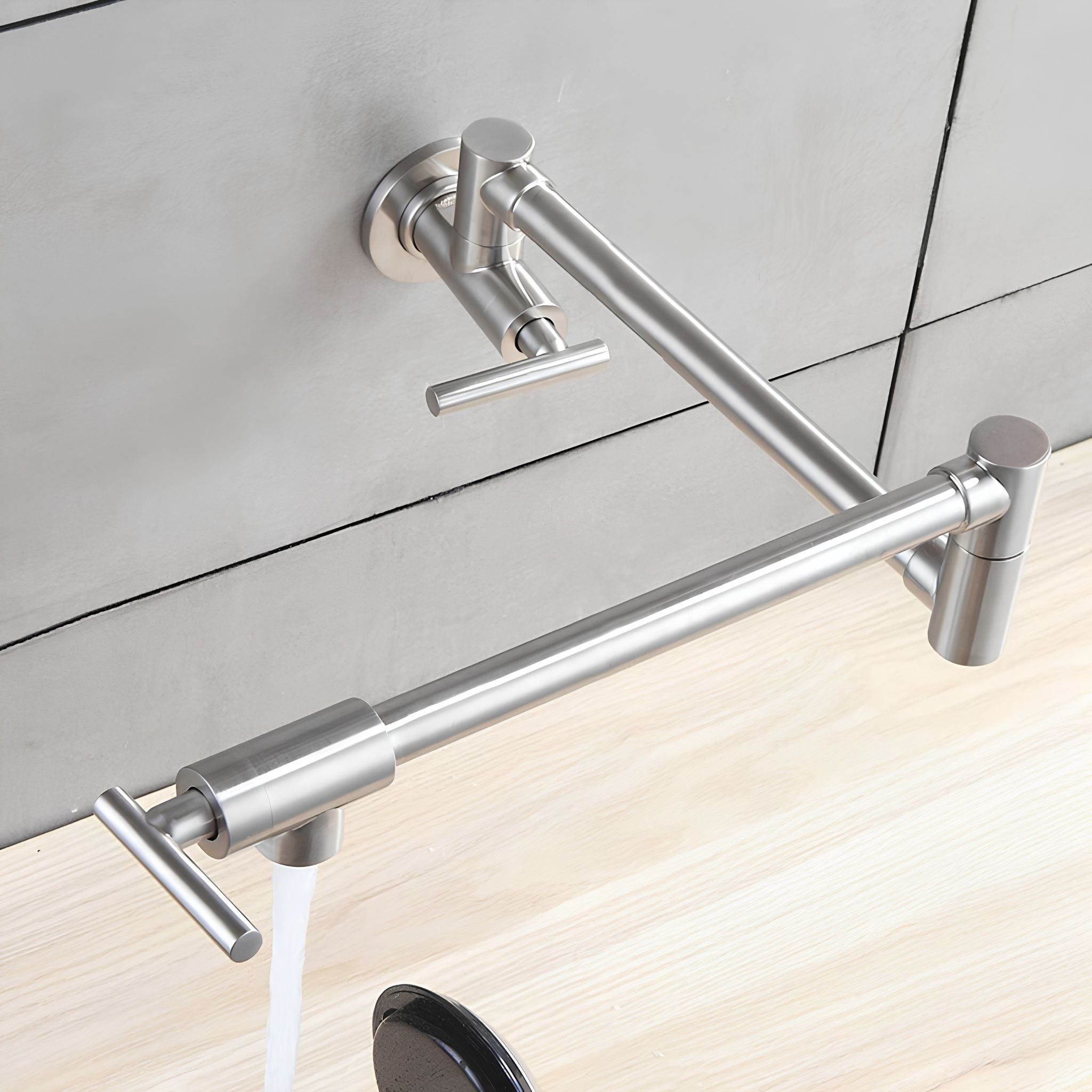 A wall-mounted stainless steel faucet with a curved spout and side lever handle, shown with water flowing from the spout onto a light wood surface against a gray tiled wall. #color_brushed nickel