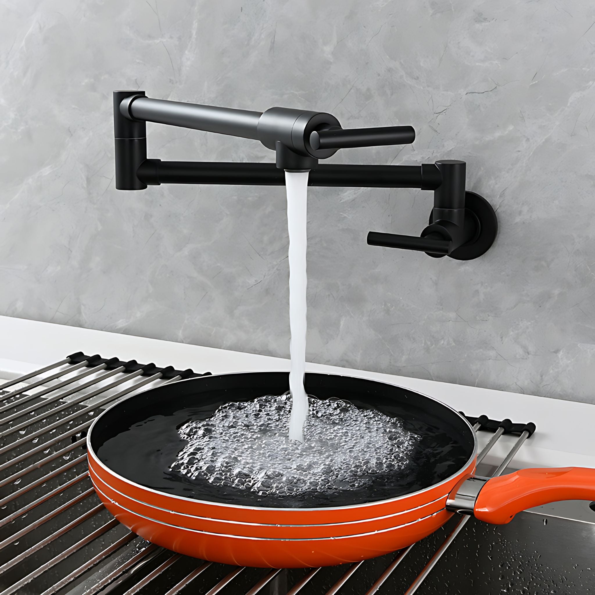 An orange non-stick frying pan sits on a metal sink rack beneath a matte black wall-mounted faucet with water running into the pan, set against a gray stone backsplash. #color_matte black