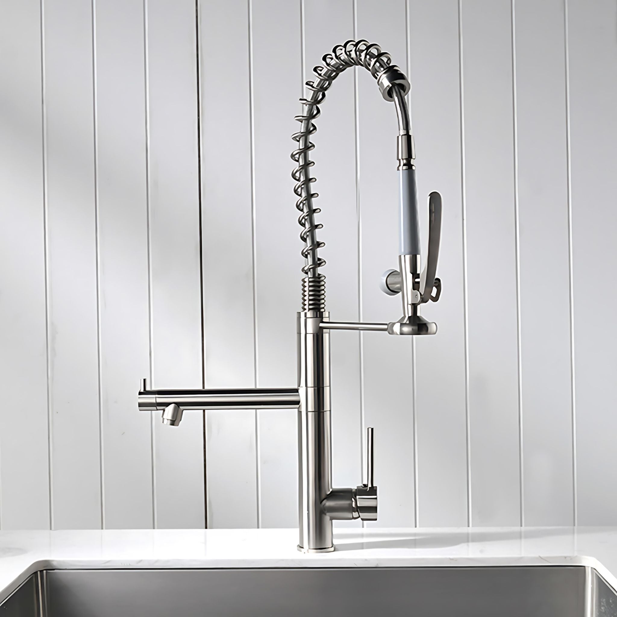 A modern pull-down kitchen faucet with a flexible spring-coil hose, single lever handle, and side spout, shown installed above a sink against a white paneled wall. #color_brushed nickel