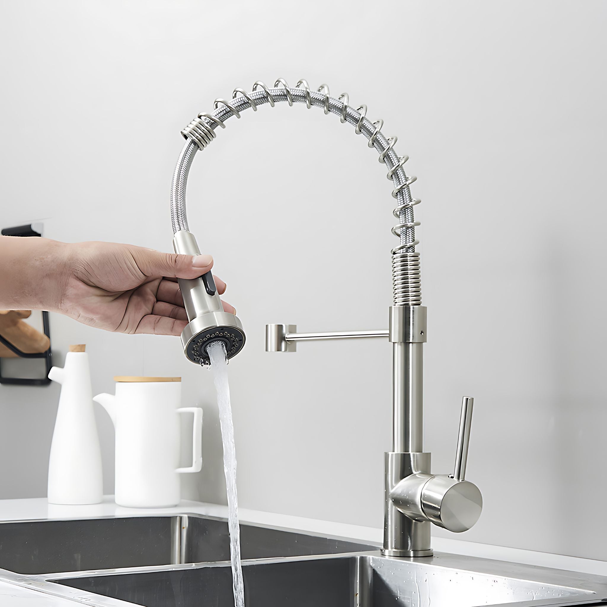 A modern pull-down kitchen faucet with a flexible gooseneck spout in brushed silver stainless steel, shown with a hand holding the spray head under running water in a stainless steel sink. #color_brushed nickel