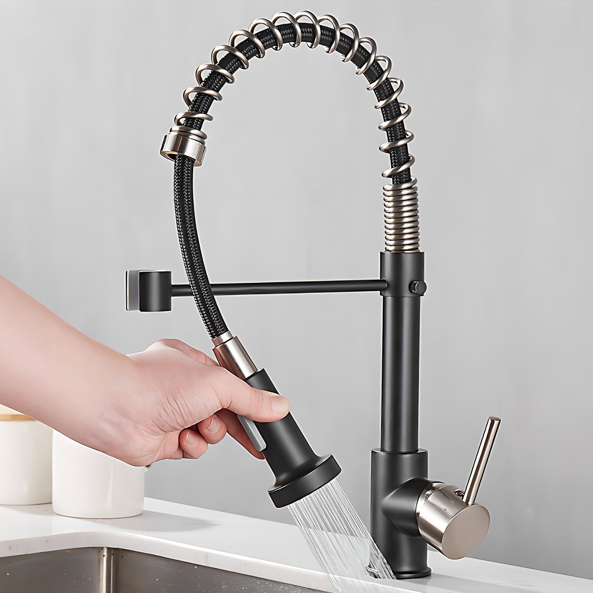 A modern kitchen faucet with a flexible gooseneck spout and side-mounted spray head, shown with a hand holding the sprayer over running water. The faucet has a matte black body with brushed silver metal accents, installed on a white countertop above a sink. #color_matte black and brushed nickel
