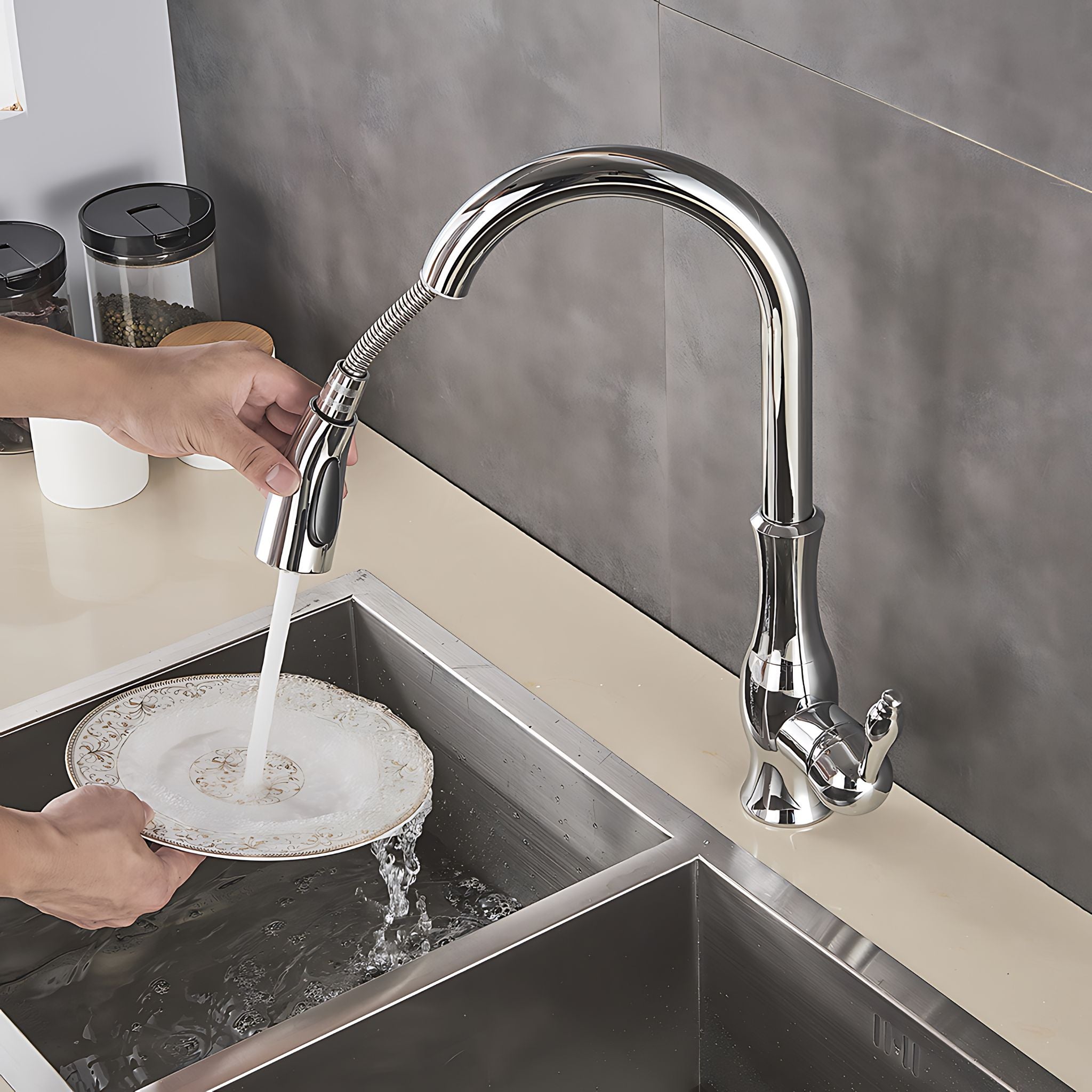 A polished chrome pull-down kitchen faucet with running water is shown being used to rinse a plate in a stainless steel double sink, with a person's hands visible. #color_chrome