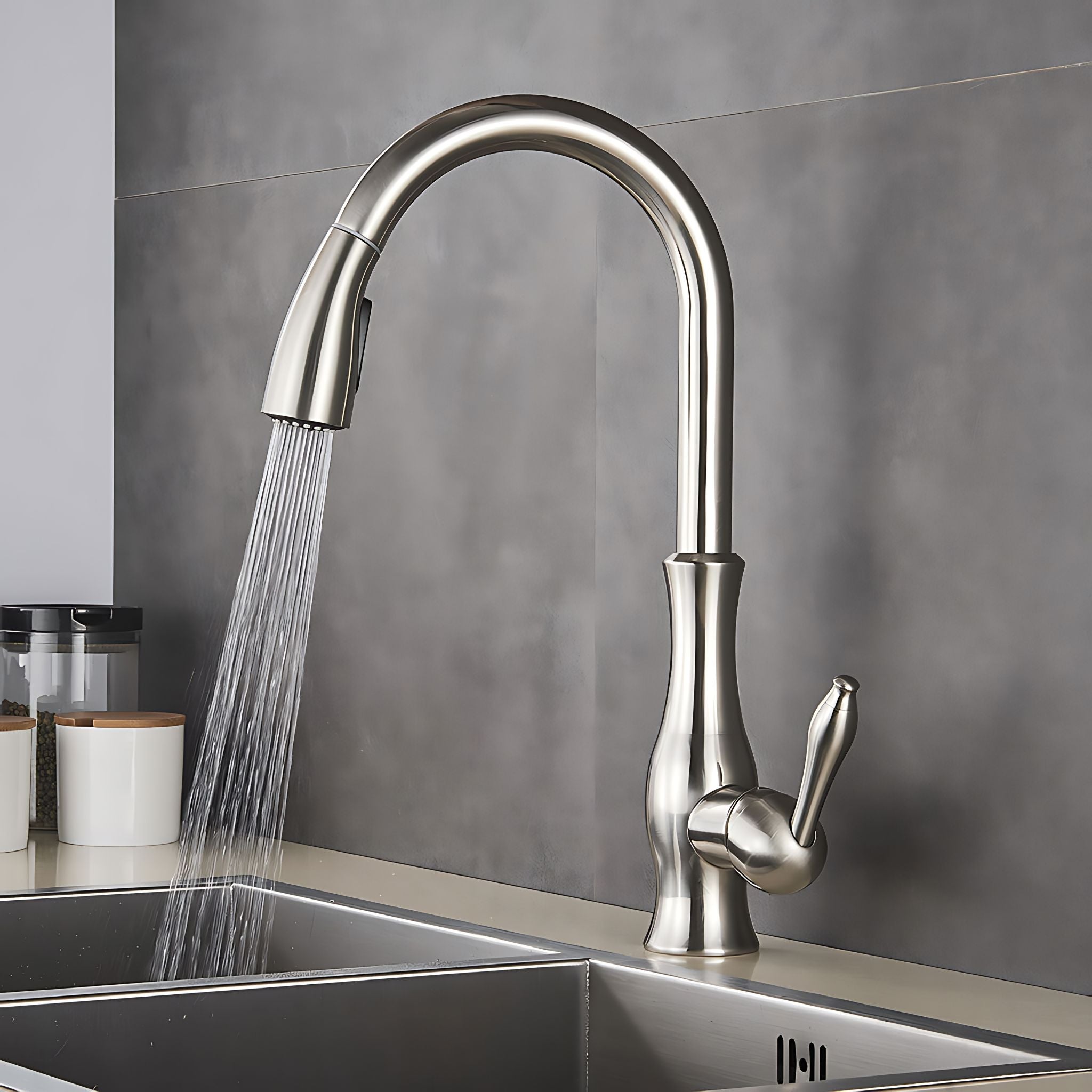 A modern pull-down kitchen faucet with a curved high-arc spout and single lever handle, shown with water running into a double-basin sink against a gray tiled backsplash. #color_brushed nickel