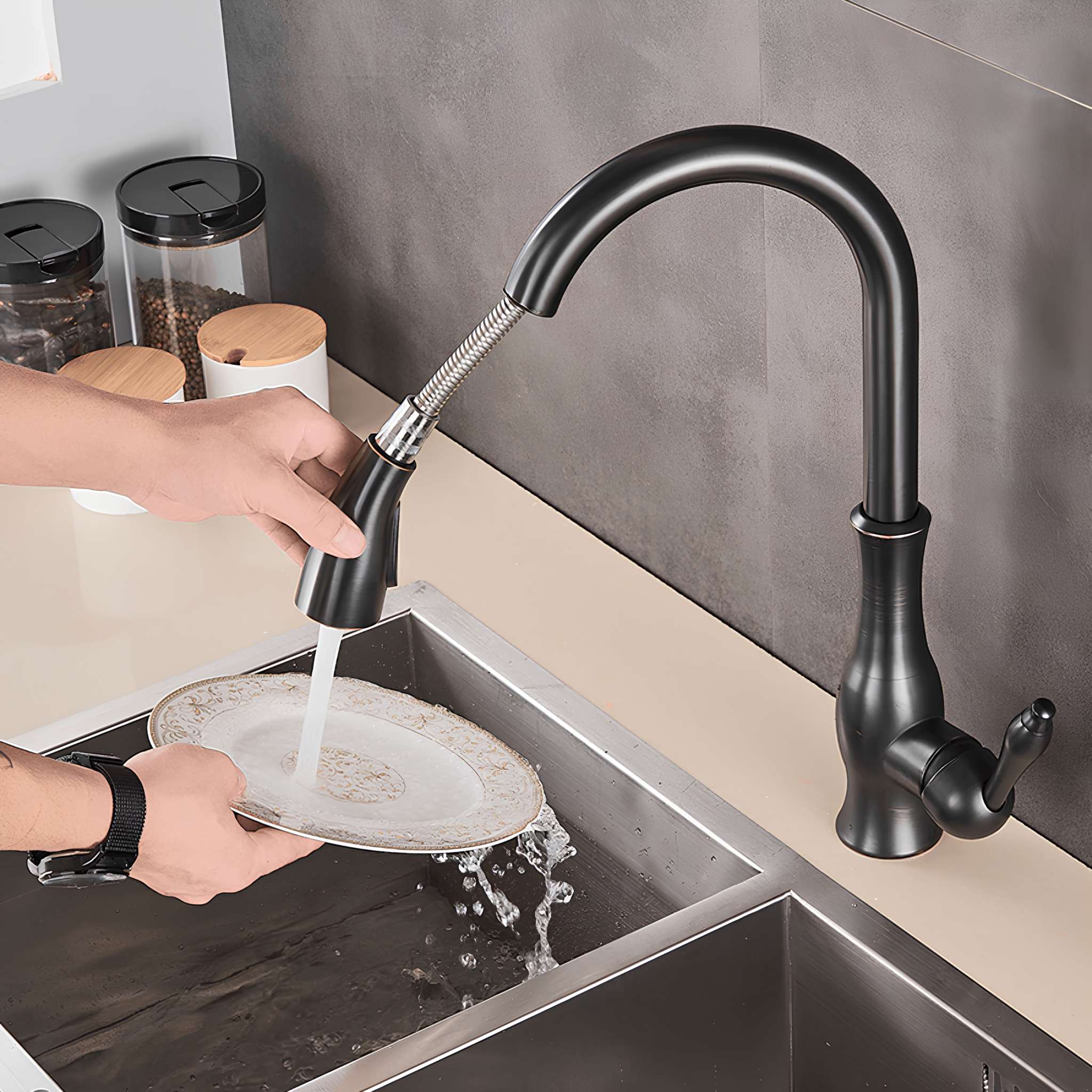 A matte black pull-down kitchen faucet with a flexible spring hose is shown running water while a person washes a plate in a stainless steel sink. The faucet is installed on a beige countertop against a gray tiled backsplash. #color_matte black