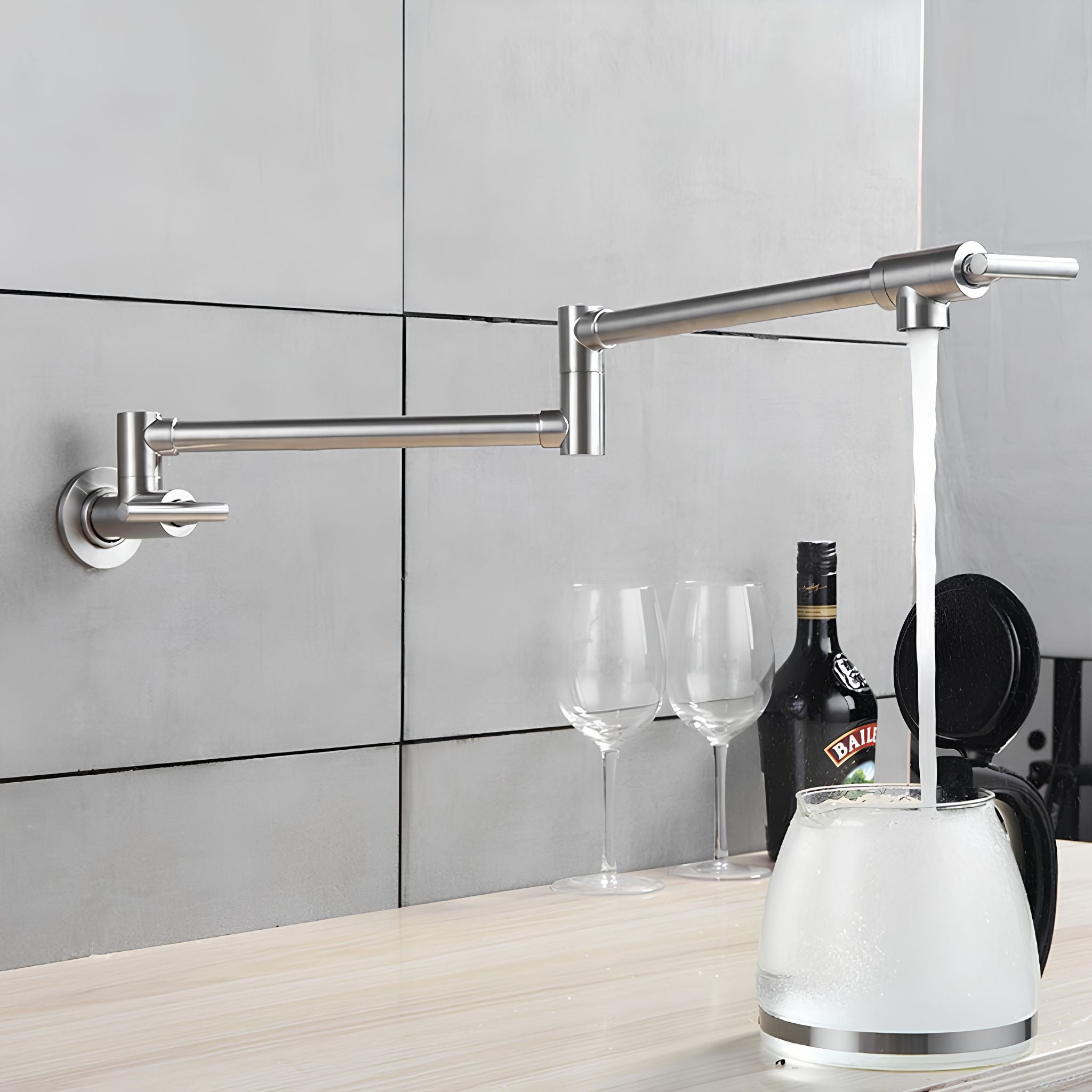 A wall-mounted stainless steel kitchen faucet with an adjustable arm, shown with water running into a white electric kettle on a light wood countertop. Two clear wine glasses and a dark bottle are visible against a gray tiled backsplash. #color_brushed nickel