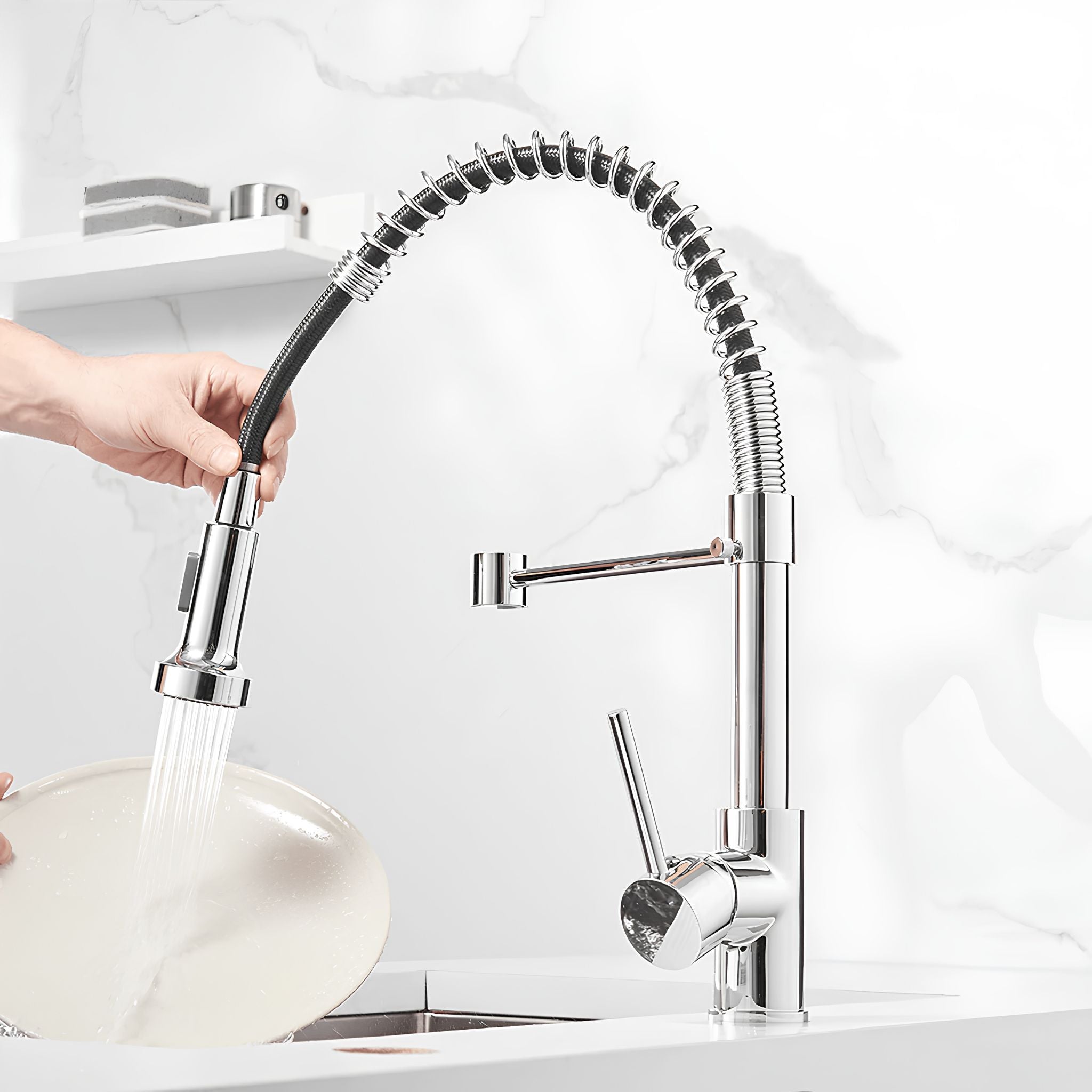 A high-arc chrome kitchen faucet with a flexible pull-out sprayer, shown with a hand turning it on and water flowing. The faucet is installed on a white sink with a white marble backsplash, and a hand is holding a plate under the running water. #color_chrome