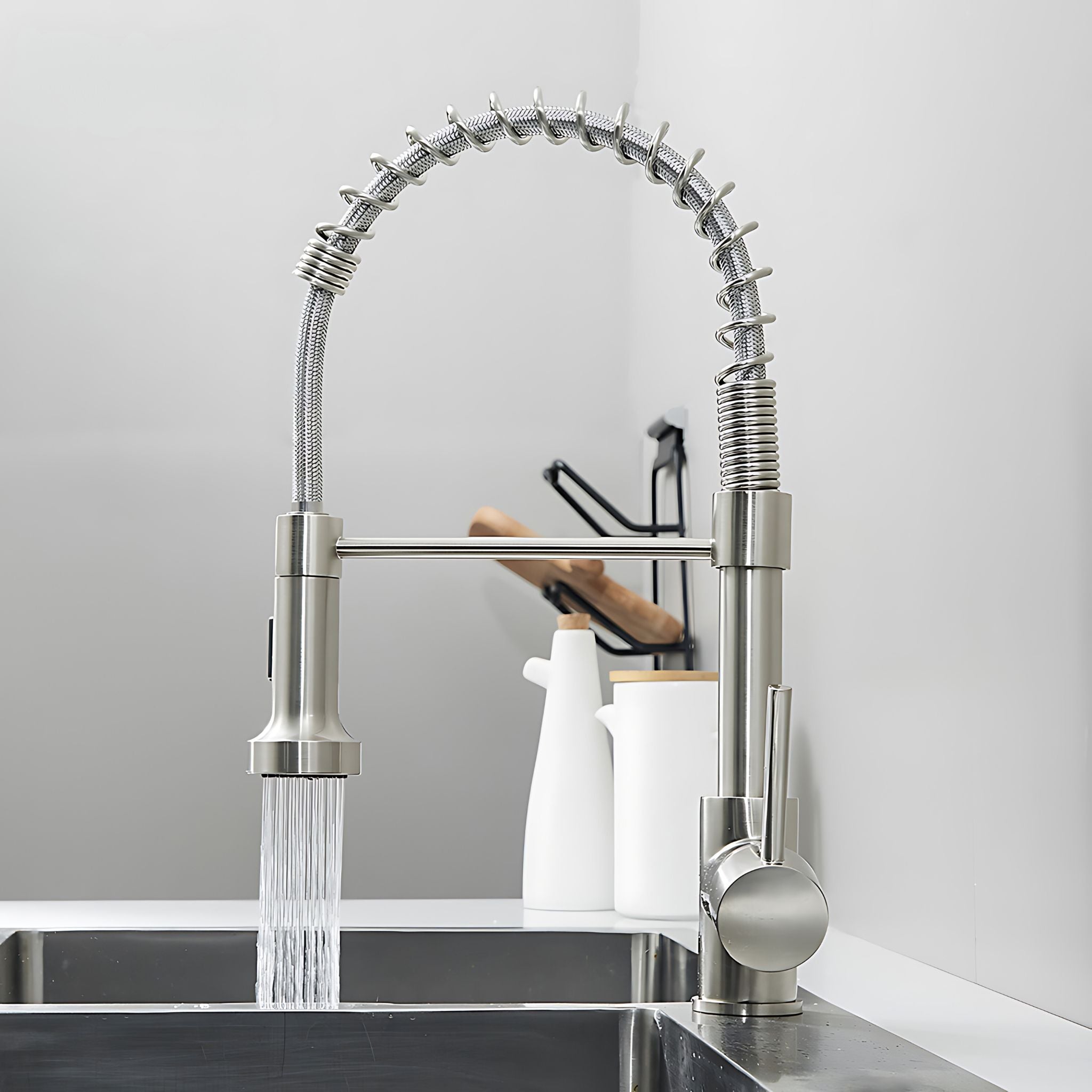 A modern pull-down faucet with a flexible high-arc spout in brushed stainless steel, shown installed over a sink with water running. The faucet has a single round handle and is set against a clean white kitchen wall with white ceramic containers in the background. #color_brushed nickel
