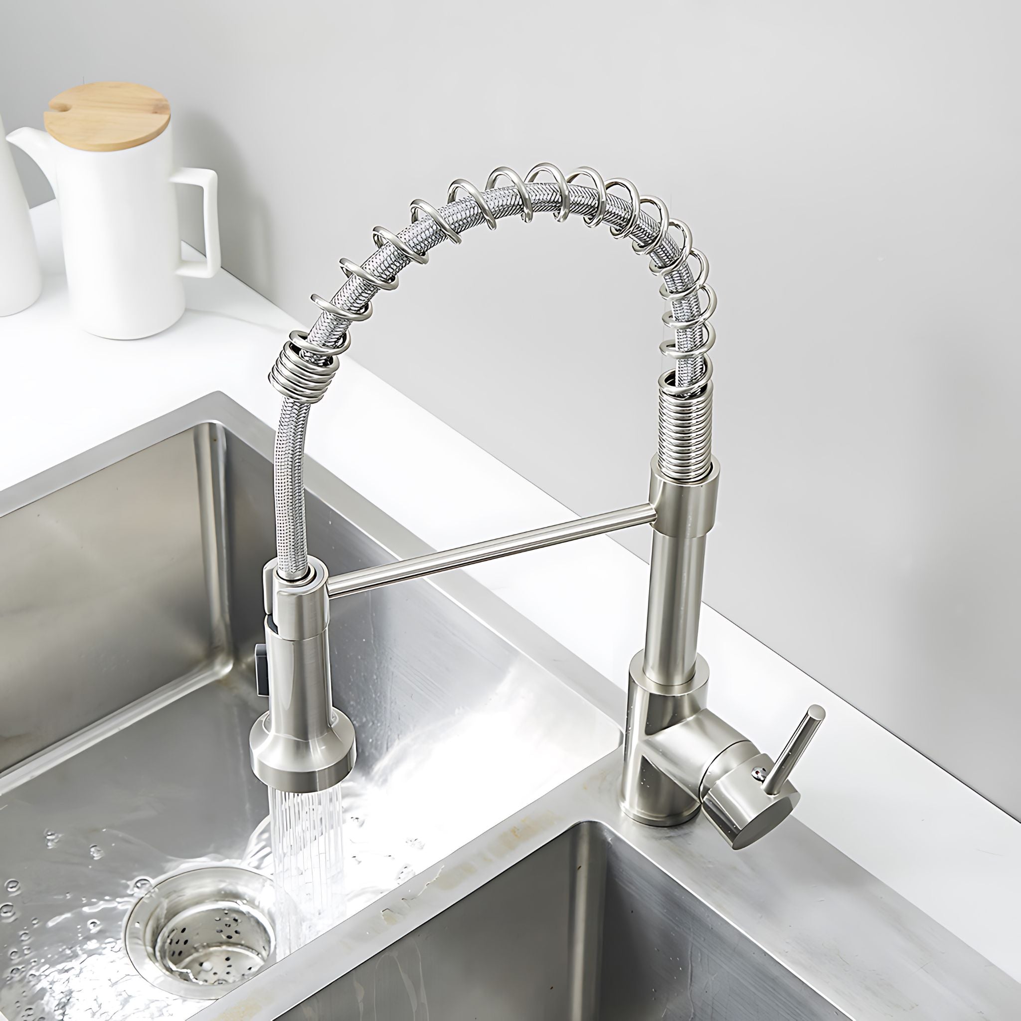 A modern stainless steel pull-down kitchen faucet with a flexible coiled hose, shown running over a double-basin sink with water flowing. The faucet has a brushed silver finish and is installed on a white countertop in a clean kitchen setting. #color_brushed nickel