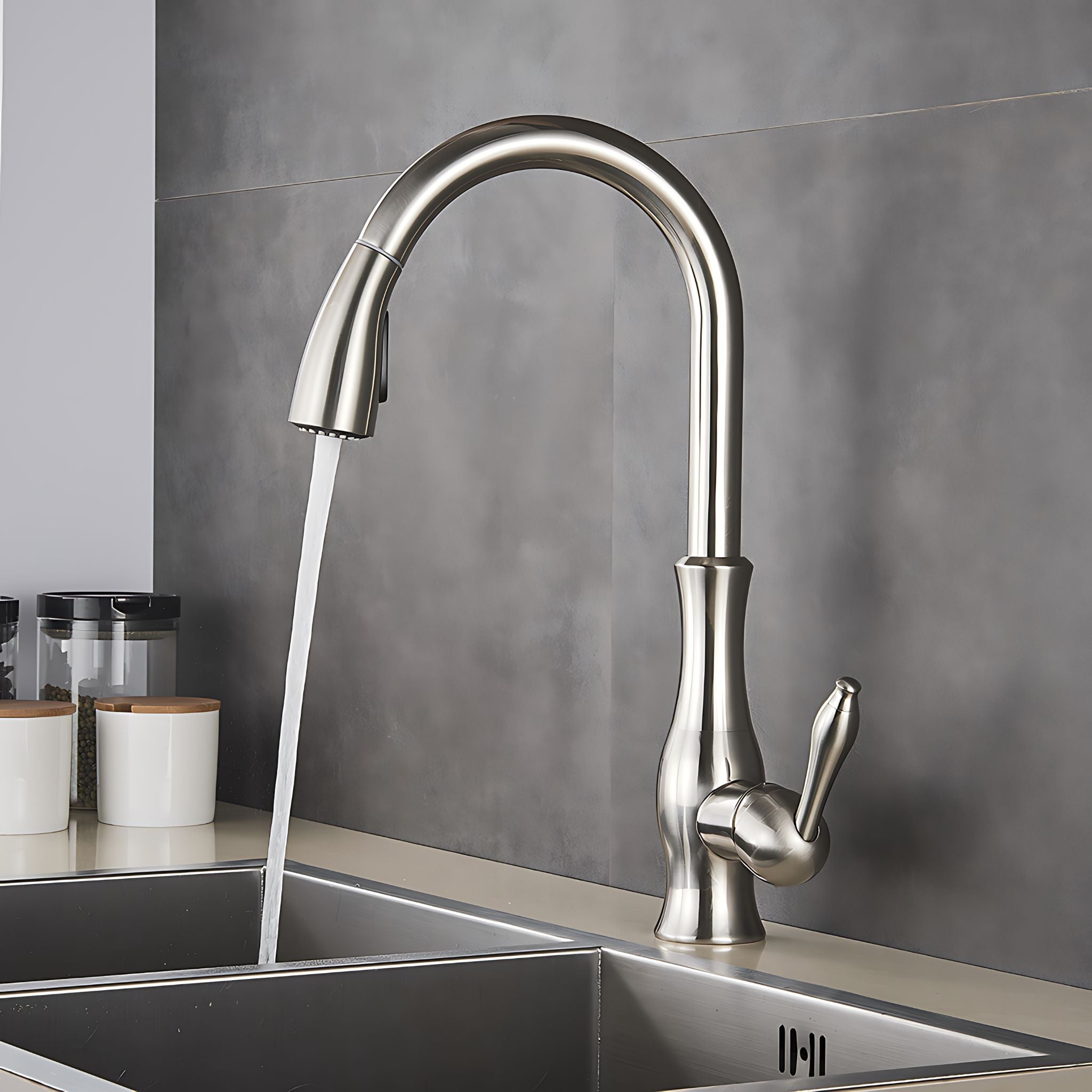 A modern pull-down kitchen faucet with a curved high-arc spout and single lever handle, shown running water into a stainless steel double sink against a gray tiled backsplash. #color_brushed nickel