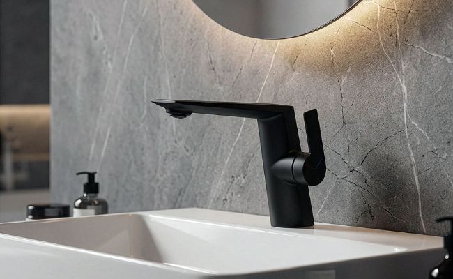 Kissinger modern single-hole bathroom sink faucet in a matte black finish, featuring a sleek side single-handle design, installed on a white basin sink against a gray marble wall beneath a backlit round mirror.