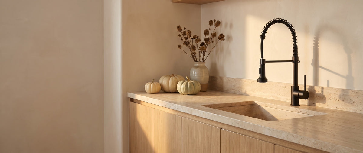 Matte black Spiro spring spout kitchen faucet at a travertine sink in a warm oak and plaster kitchen with autumn accents, Signature Faucets Fall Sale